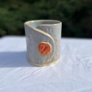 Könnte beinhalten: Handgefertigte Keramik-Tasse in Hellblau-Grau mit strukturierter Oberfläche und einem knopfartigen Detail in Rot-Orange. Die Tasse hat eine zylindrische Form mit einem gebogenen Griff. Geeignet für Heiß- oder Kaltgetränke.