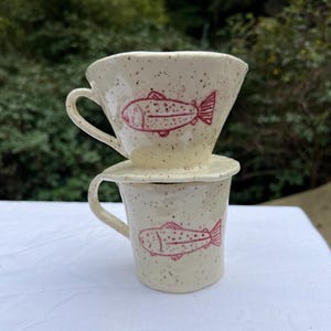 May include: A cream-coloured ceramic coffee dripper and mug set. Each piece features a hand-drawn red fish design. The set is speckled with dark brown spots. The dripper sits atop the mug, ready for brewing.