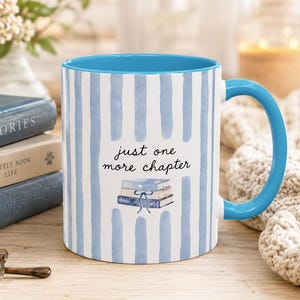May include: A white ceramic mug with a bright blue handle and interior, featuring vertical light blue stripes. The front displays a stack of three illustrated books with a blue ribbon and the black text "just one more chapter" centered.