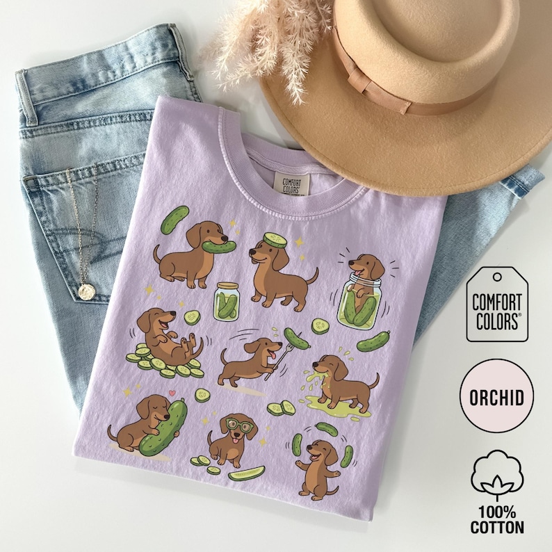 May include: Orchid-colored t-shirt featuring a repeating pattern of cartoon dachshunds interacting with pickles. The Comfort Colors shirt is made of 100% cotton. A pair of blue jeans and a tan hat are in the background.