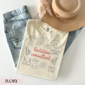 May include: Ivory-colored t-shirt with a "lactation consultant" design in red and blue. The shirt is laid on top of light blue jeans, a gold necklace, and a tan hat. The design includes illustrations of breastfeeding tools and a bra.