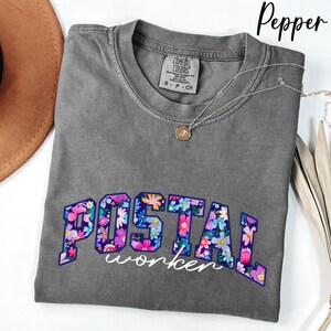May include: A grey Comfort Colors t-shirt with the words "POSTAL worker" in a floral design. A silver necklace with a pendant is draped across the shirt. The shirt is folded on a white surface.