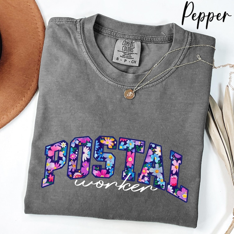 May include: A grey Comfort Colors t-shirt with the words "POSTAL worker" in a floral design. A silver necklace with a pendant is draped across the shirt. The shirt is folded on a white surface.