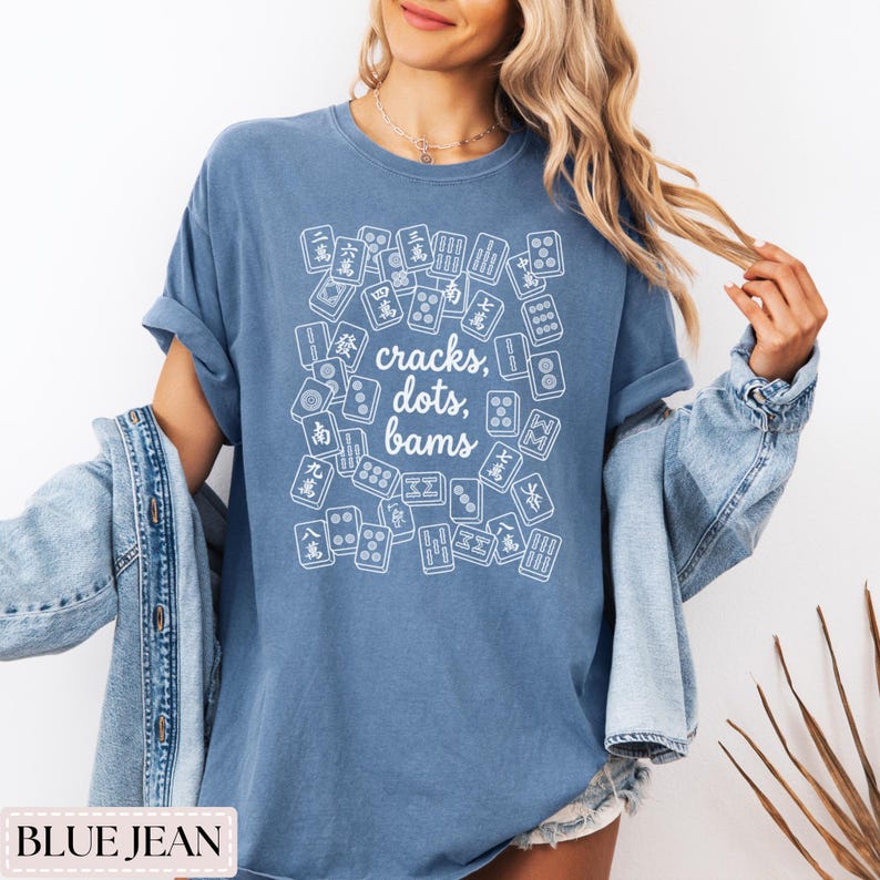 May include: Blue jean colored t-shirt with a white graphic of Mahjong tiles and the text "cracks, dots, bams". The shirt has short sleeves and a relaxed fit. The shirt is made of a soft material.