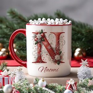 Op de afbeelding: Een witte keramische mok met een rode handgreep en rand, gevuld met marshmallows. De voorzijde toont een grote rode letter N versierd met hulst, bessen en sneeuwvlokken, met de naam Naomi in een rood cursief lettertype eronder.