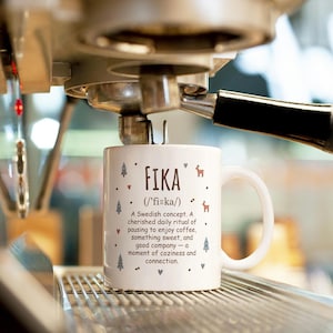 May include: White ceramic mug with the word "FIKA" in black, accompanied by a definition and illustrations of trees, hearts, and reindeer. The mug is on a coffee machine.