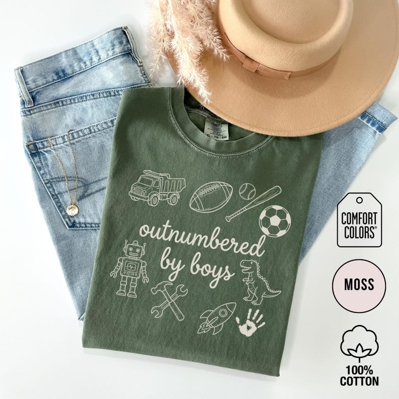 May include: A moss green t-shirt with the phrase "outnumbered by boys" and various white graphics, including a truck, football, baseball, soccer ball, robot, hammer, wrench, rocket, dinosaur, and handprint. The shirt is paired with light blue jeans and a tan hat.