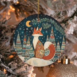 Op de afbeelding: Ronde kerstversiering met een vos in een winterbos. De vos heeft oranje en witte vacht, met een patroon op zijn borst. De achtergrond is een donkerblauwe nachthemel met een maan, sneeuwvlokken en versierde bomen.