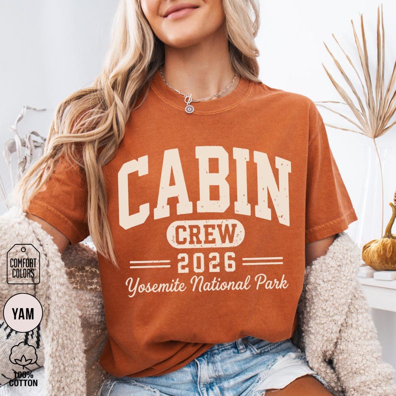 May include: A burnt orange short-sleeve t-shirt featuring cream-colored distressed text that reads CABIN CREW 2026 Yosemite National Park. The shirt has a crew neckline and a relaxed fit, with a graphic design centered on the front chest area.