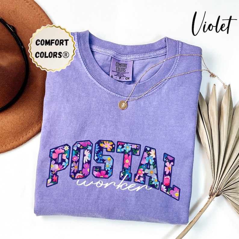 May include: A violet Comfort Colors t-shirt featuring the words "POSTAL worker" in a floral design. The shirt has a crew neck and short sleeves. A gold necklace with a pendant is draped on the shirt. A brown hat and dried plant are in the background.