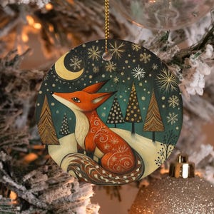 May include: A round ceramic Christmas ornament featuring a painted orange fox sitting in a snowy landscape with stylized evergreen trees. The dark teal night sky is decorated with a golden crescent moon and white snowflake patterns.