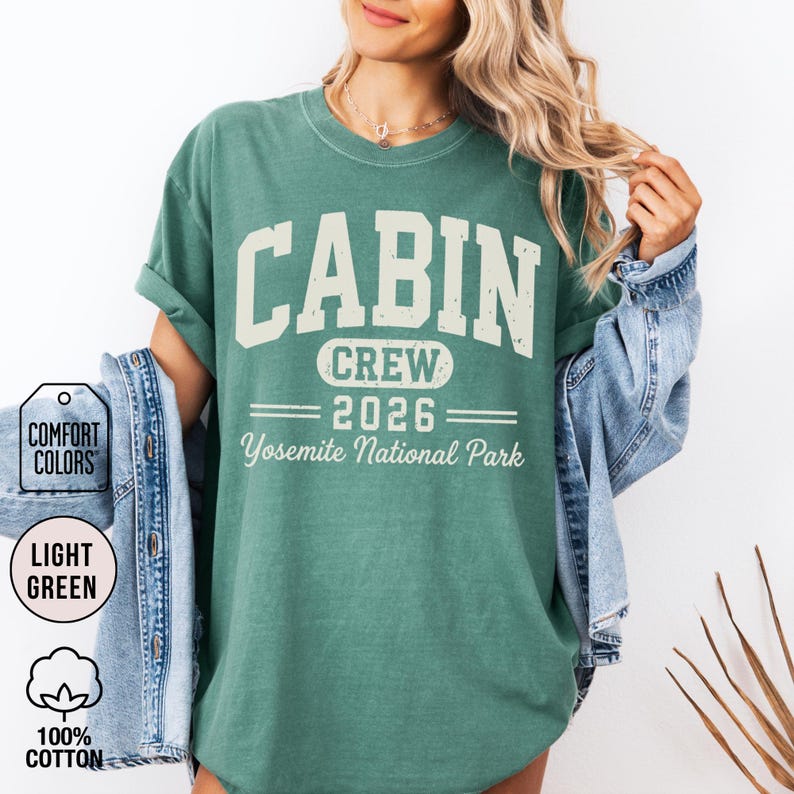 May include: A light green short-sleeve t-shirt featuring the text CABIN CREW 2026 Yosemite National Park in a distressed white font. The shirt is layered with a blue denim jacket. Two circular labels indicate Comfort Colors and 100% cotton.