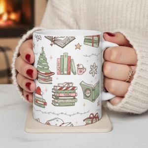 May include: White ceramic mug featuring a repeating pattern of books, Christmas trees, snowflakes, and gifts in shades of green, red, and pink. The mug has a white handle and is sitting on a beige coaster.