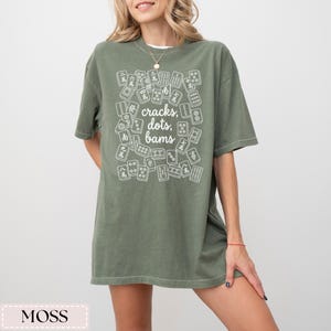 May include: Moss green t-shirt with a white graphic of Mahjong tiles and the text "cracks, dots, bams." The shirt is a relaxed fit and has short sleeves. The shirt is made of a soft material.