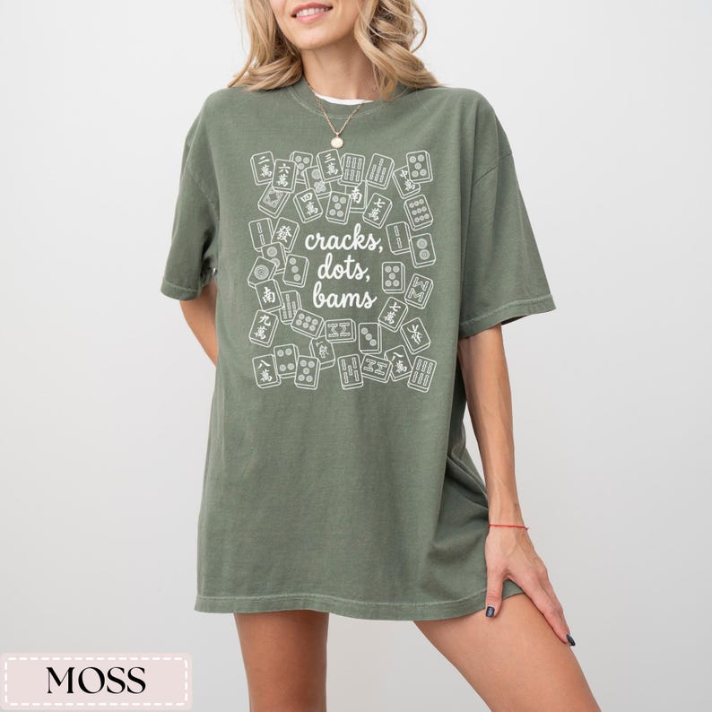 May include: Moss green t-shirt with a white graphic of Mahjong tiles and the text "cracks, dots, bams." The shirt is a relaxed fit and has short sleeves. The shirt is made of a soft material.