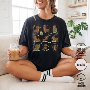 May include: Black t-shirt with a repeating pattern of dachshunds and pickles. The dachshunds are brown and the pickles are green. The shirt has the words "Comfort Colors", "Black", and "100% Cotton" printed on it.