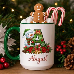 May include: A white ceramic mug with a green handle and rim, decorated with a snowman, the letter "A", and the name "Abigail". The mug is filled with marshmallows, a gingerbread man, and candy canes. Festive holiday theme.