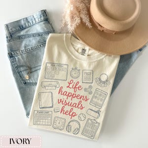 May include: Ivory-colored t-shirt with the phrase "Life happens visuals help" in red script. The shirt features various visual aids in blue, including a visual schedule, timer, and reward chart. A light brown hat and light blue jeans are also in the image.