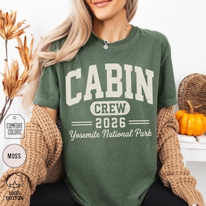 May include: A moss green short-sleeve t-shirt featuring the text CABIN CREW 2026 Yosemite National Park in a distressed cream-colored font. The shirt has a crew neckline and is layered under a tan knit cardigan, showcasing a casual style.