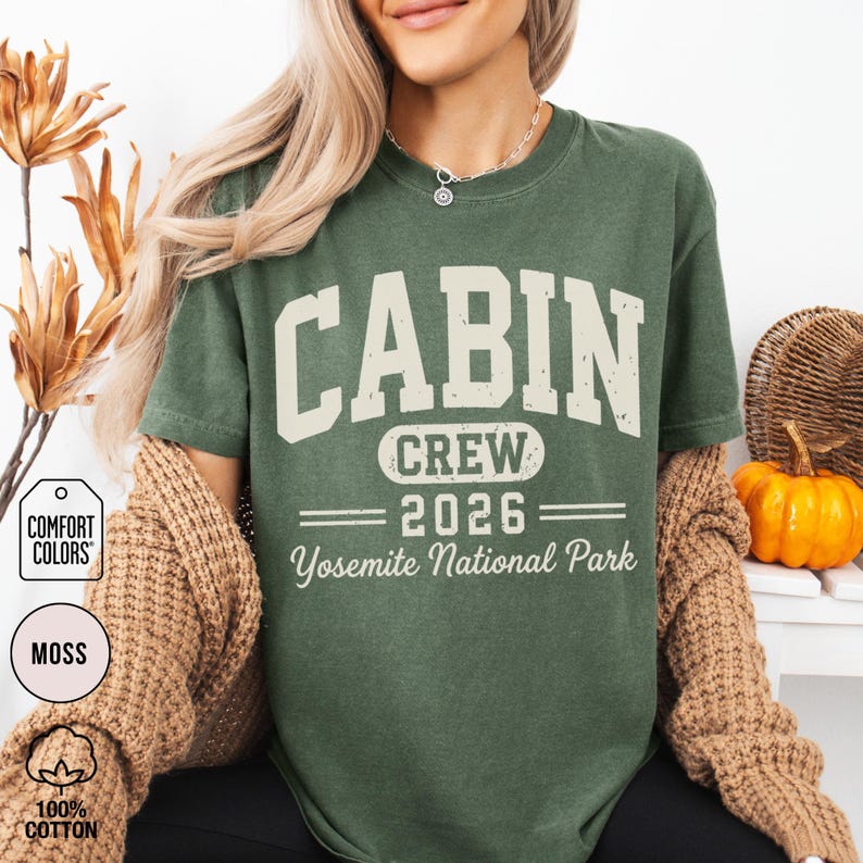 May include: A moss green short-sleeve t-shirt featuring the text CABIN CREW 2026 Yosemite National Park in a distressed cream-colored font. The shirt has a crew neckline and is layered under a tan knit cardigan, showcasing a casual style.