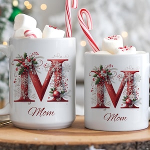 Op de afbeelding: Twee witte keramische mokken met een grote rode letter M versierd met hulst, bessen en sneeuwvlokken. Onder de letter staat het woord Mom in cursief schrift gedrukt. Elke mok is gevuld met witte marshmallows en een rood-witte zuurstok.