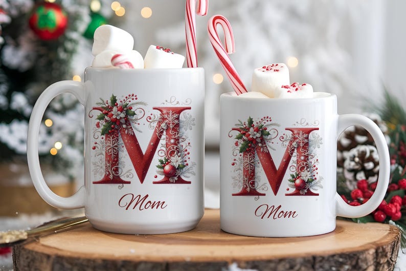 Op de afbeelding: Twee witte keramische mokken met een grote rode letter M versierd met hulst, bessen en sneeuwvlokken. Onder de letter staat het woord Mom in cursief schrift gedrukt. Elke mok is gevuld met witte marshmallows en een rood-witte zuurstok.