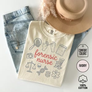 May include: Ivory-colored t-shirt with the words "forensic nurse" in red script, surrounded by forensic science-themed illustrations. The shirt is paired with light-wash jeans, a necklace, and a beige hat. The shirt is made of 100% cotton.