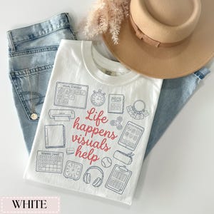 May include: White t-shirt with a crew neck featuring a graphic design with the text "Life happens visuals help" in red and blue. The shirt is paired with light blue jeans and a tan hat.