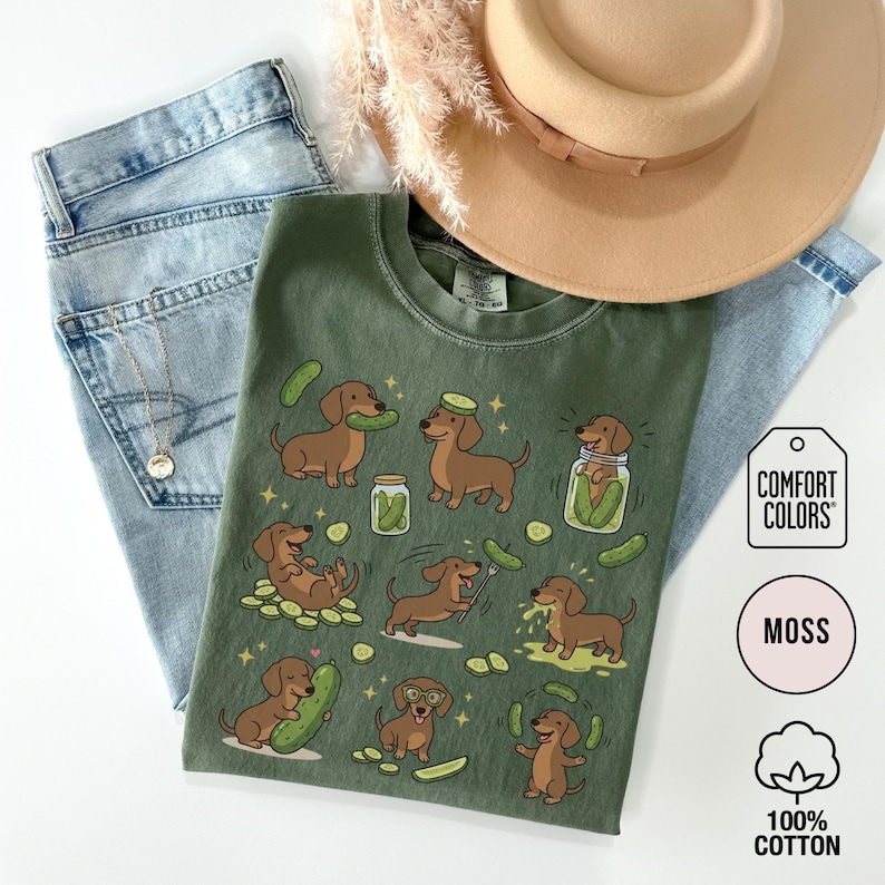May include: A moss green Comfort Colors t-shirt featuring a playful design of dachshunds interacting with pickles. The dogs are depicted with pickles, in jars, and wearing pickle hats. The shirt is made of 100% cotton and is paired with blue jeans and a tan hat.