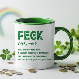 May include: White ceramic mug with a green rim and handle. The mug features a green graphic with the word "FECK" in large letters and the definition "(fek) verb" underneath. The text continues with "IRELAND'S MOST USED WORD. A UNIQUELY IRISH WAY OF EXPRESSING ONE'S ANNOYANCE OR FRUSTRATION. See also: feck off, feck sake, feckin eejit"