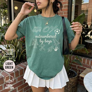 May include: Light green t-shirt with the text "outnumbered by boys" and various white graphic illustrations. The graphics include a truck, football, baseball, soccer ball, robot, wrench, rocket, dinosaur, and handprint. The shirt is made of 100% cotton.
