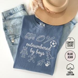 May include: A blue Comfort Colors t-shirt with the phrase "outnumbered by boys" and various white graphics, including a truck, football, baseball, soccer ball, robot, and dinosaur. The shirt is paired with blue jeans and a tan hat.