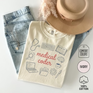May include: Ivory-colored t-shirt with the words "medical coder" in red script, surrounded by medical-related icons. The shirt is made of 100% cotton and is paired with light-wash jeans and a tan hat. The shirt also has the Comfort Colors logo.