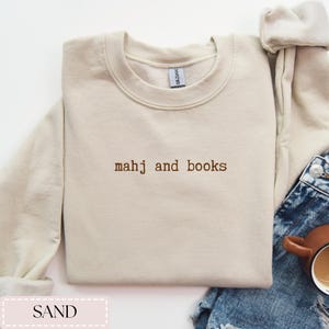 May include: Sand-colored crewneck sweatshirt with the embroidered text "mahj and books" in brown. The sweatshirt is made of a soft material and has a classic, casual design. The image also shows a portion of blue denim jeans.