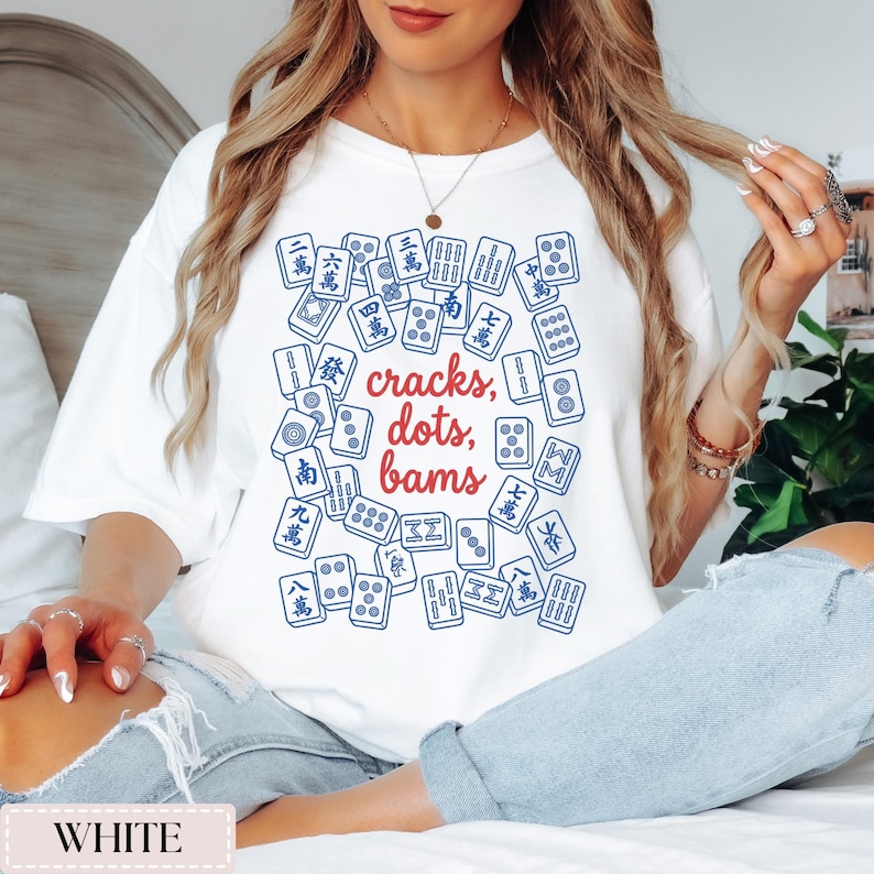 May include: White t-shirt featuring a design of blue and red Mahjong tiles. The text "cracks, dots, bams" is in the center. The shirt is a casual style.