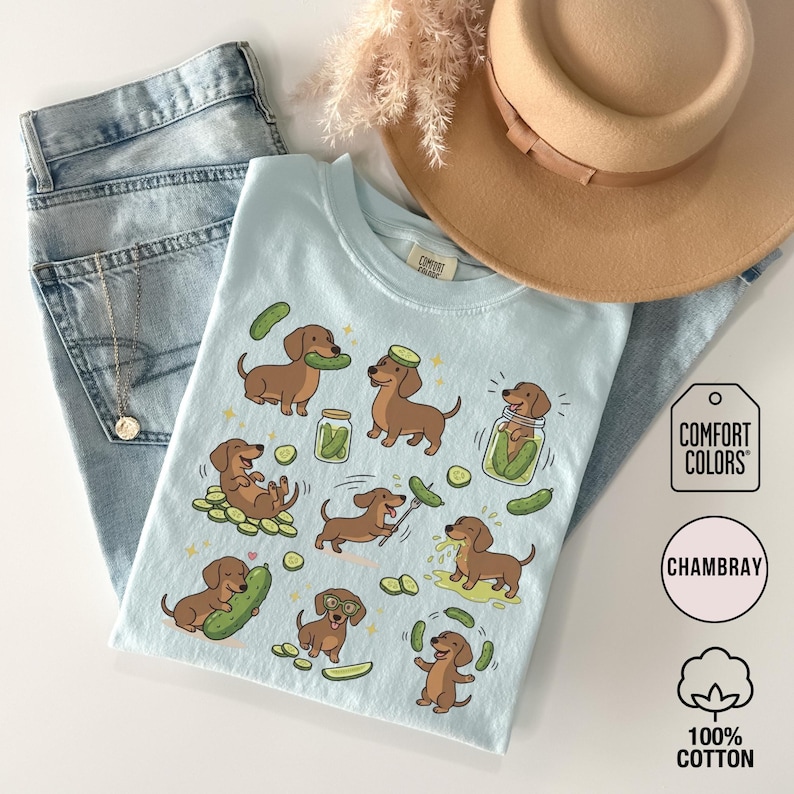 May include: A light blue t-shirt featuring cartoon dachshunds interacting with pickles. The shirt is paired with light wash jeans and a tan hat. The shirt has the words "Comfort Colors", "Chambray", and "100% Cotton" printed on it.