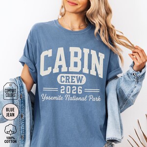 May include: A blue jean colored short-sleeve t-shirt featuring the text CABIN CREW 2026 Yosemite National Park in a distressed white font. The shirt has a crew neckline and is layered under a denim jacket. A Comfort Colors brand tag is visible.