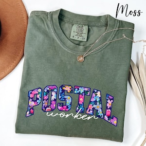 May include: A moss green t-shirt featuring the text "POSTAL worker" in a floral pattern. The shirt has a crew neck and short sleeves. A silver necklace with a small pendant is draped on the shirt.