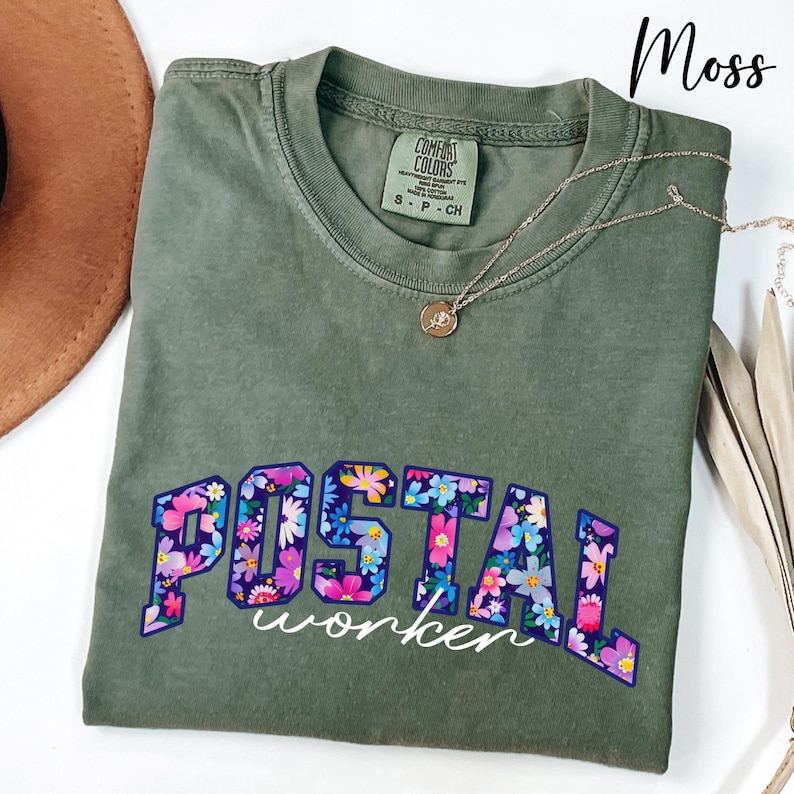 May include: A moss green t-shirt featuring the text "POSTAL worker" in a floral pattern. The shirt has a crew neck and short sleeves. A silver necklace with a small pendant is draped on the shirt.