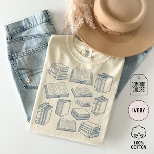 May include: An ivory t-shirt featuring a blue line-art graphic of various stacked and open books. The shirt is paired with light blue denim jeans, a tan wide-brimmed hat, and a delicate silver necklace. The Comfort Colors brand tag is visible.