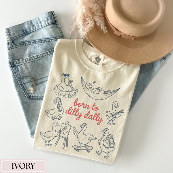 May include: Ivory-colored t-shirt with a graphic of ducks in various leisurely activities, including floating on a tube, relaxing in a hammock, and playing guitar. The phrase "born to dilly dally" is printed in red. The shirt is paired with light-wash jeans and a tan hat.