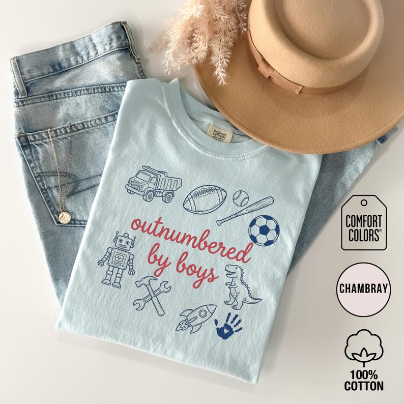 May include: A light blue t-shirt with the phrase "outnumbered by boys" in red, surrounded by illustrations of a truck, football, baseball, robot, hammer, rocket, dinosaur, soccer ball, and handprint. The shirt is paired with light wash jeans and a tan hat.