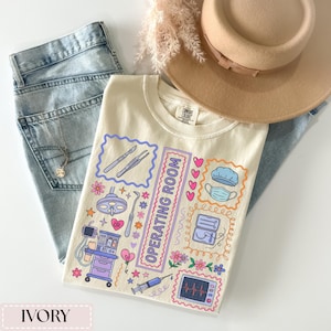 May include: Ivory-colored t-shirt with a colorful "Operating Room" graphic. The design includes medical tools, a surgical lamp, a heart monitor, and other medical-themed illustrations. The shirt is laid on top of blue jeans and a tan hat.