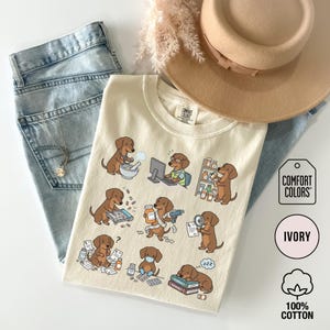 May include: An ivory t-shirt featuring a graphic print of brown dachshunds performing various pharmacy tasks, including using a computer, organizing medicine bottles, counting pills, and sleeping on books. The shirt is paired with blue jeans and a tan hat.