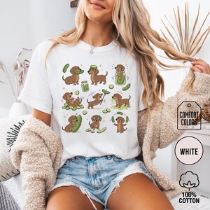 May include: White t-shirt featuring a repeating pattern of cartoon dachshunds interacting with pickles. The dachshunds are brown with various poses, and the pickles are green. The shirt has the words "COMFORT COLORS", "WHITE", and "100% COTTON" printed on it.