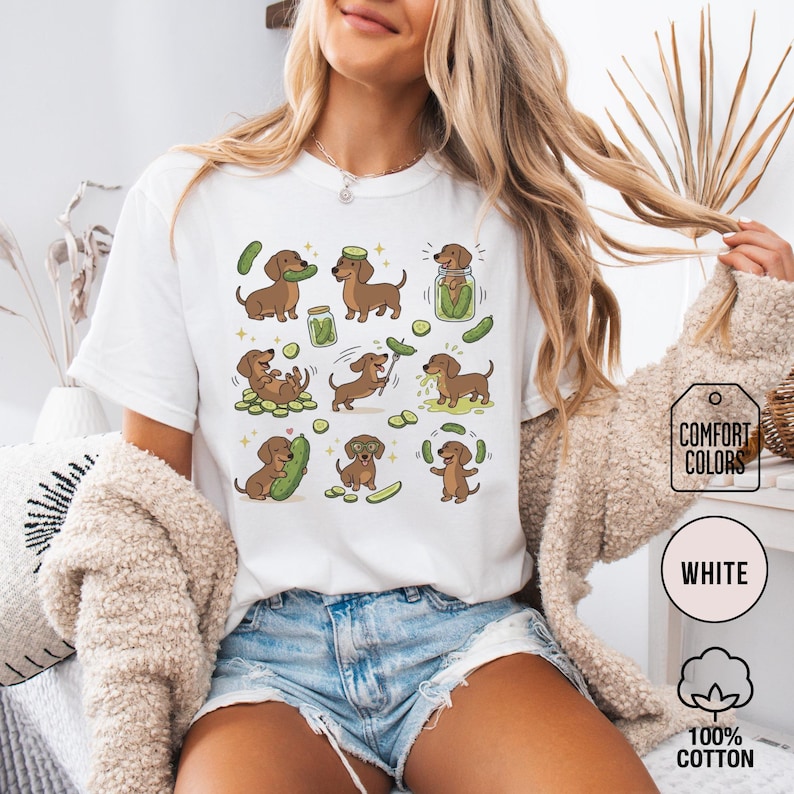 May include: White t-shirt featuring a repeating pattern of cartoon dachshunds interacting with pickles. The dachshunds are brown with various poses, and the pickles are green. The shirt has the words "COMFORT COLORS", "WHITE", and "100% COTTON" printed on it.