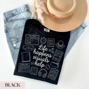 May include: Black t-shirt with the text "Life happens visuals help" and various visual aids. The shirt is laid on top of light blue jeans, a tan hat, and a sprig of dried flowers. A gold necklace is visible.