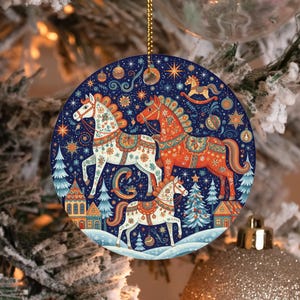 May include: A round ceramic ornament featuring a dark blue background with three horses in white and orange, decorated with intricate floral patterns. The scene includes stylized evergreen trees, small houses, stars, and ornaments in a winter setting.