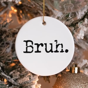 May include: A white, round Christmas ornament with the word "Bruh." printed in black, hanging from a gold string. The ornament is set against a backdrop of a decorated Christmas tree with gold and silver ornaments.