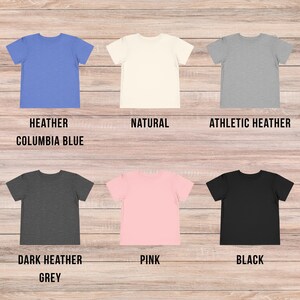 May include: Six short-sleeved t-shirts arranged in two rows. The top row features heather columbia blue, natural, and athletic heather colors. The bottom row displays dark heather grey, pink, and black colors. Each shirt has a crew neckline.